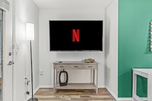 Ein Zimmer mit einem an der Wand befestigten Fernseher, der das Netflix-Logo zeigt, einem Schreibtisch mit Schubladen, einem kleinen Automodell und einer Wand mit strukturierter, grüner Oberfläche.