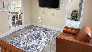 Ein Wohnzimmer mit einem braunen Ledersofa, einem gemusterten Teppich, einem an der Wand befestigten Fernseher und einer Tür, die in einen anderen Raum führt.