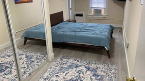Ein Schlafzimmer mit einem Bett, einem Fenster mit Jalousien, einem an der Wand montierten Fernseher und einer Tür zu einem anderen Raum.