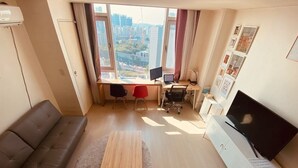 Un salon moderne comprenant un canapé, une table basse, un bureau avec deux chaises, un téléviseur et une vue sur la ville par la fenêtre.