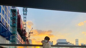 Une personne se tient sur un balcon, d’où elle a une vue sur un paysage urbain avec des bâtiments et un coucher de soleil.