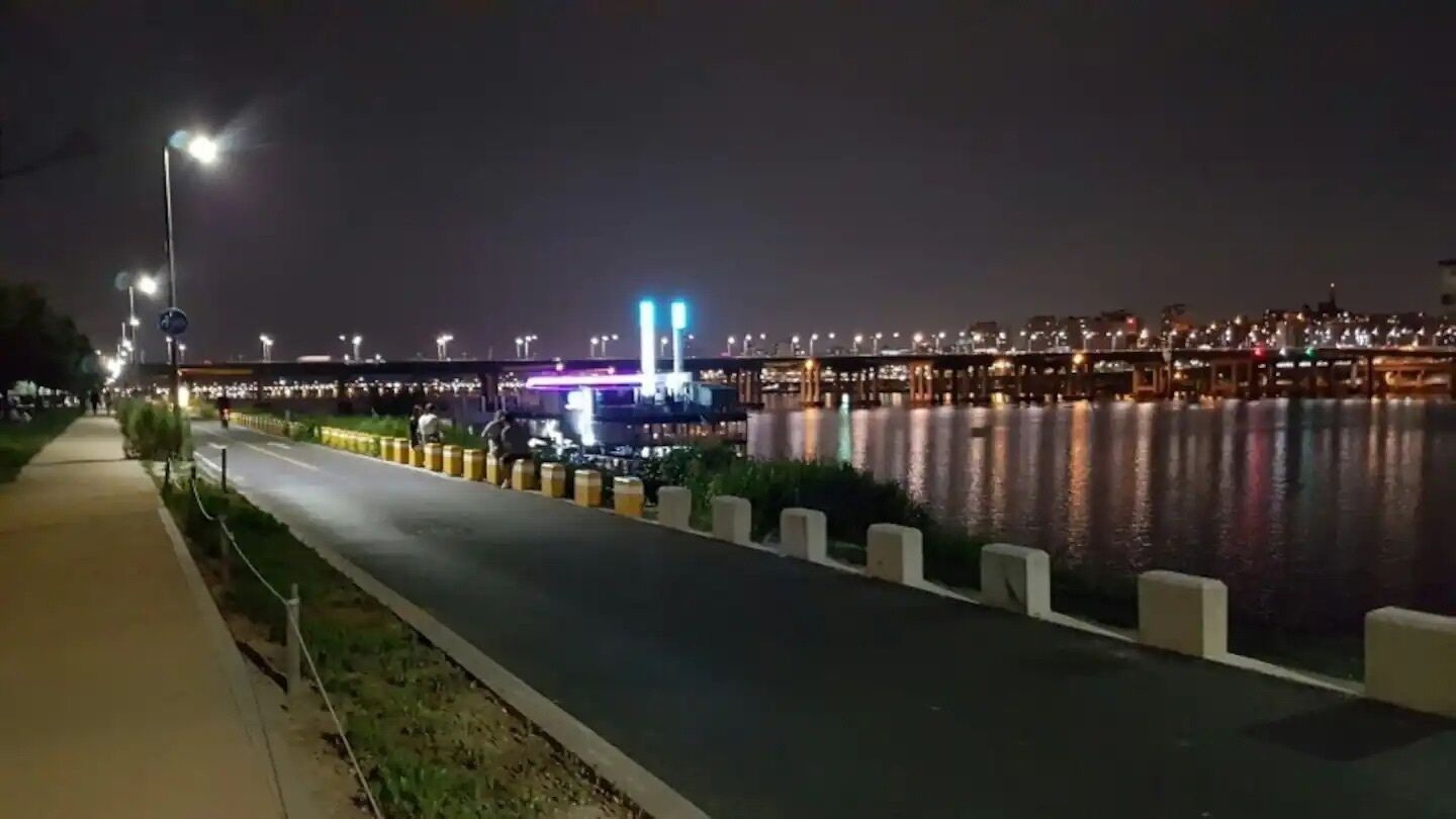 Una vista nocturna de un paseo a orillas del río, con farolas y un puente al fondo.