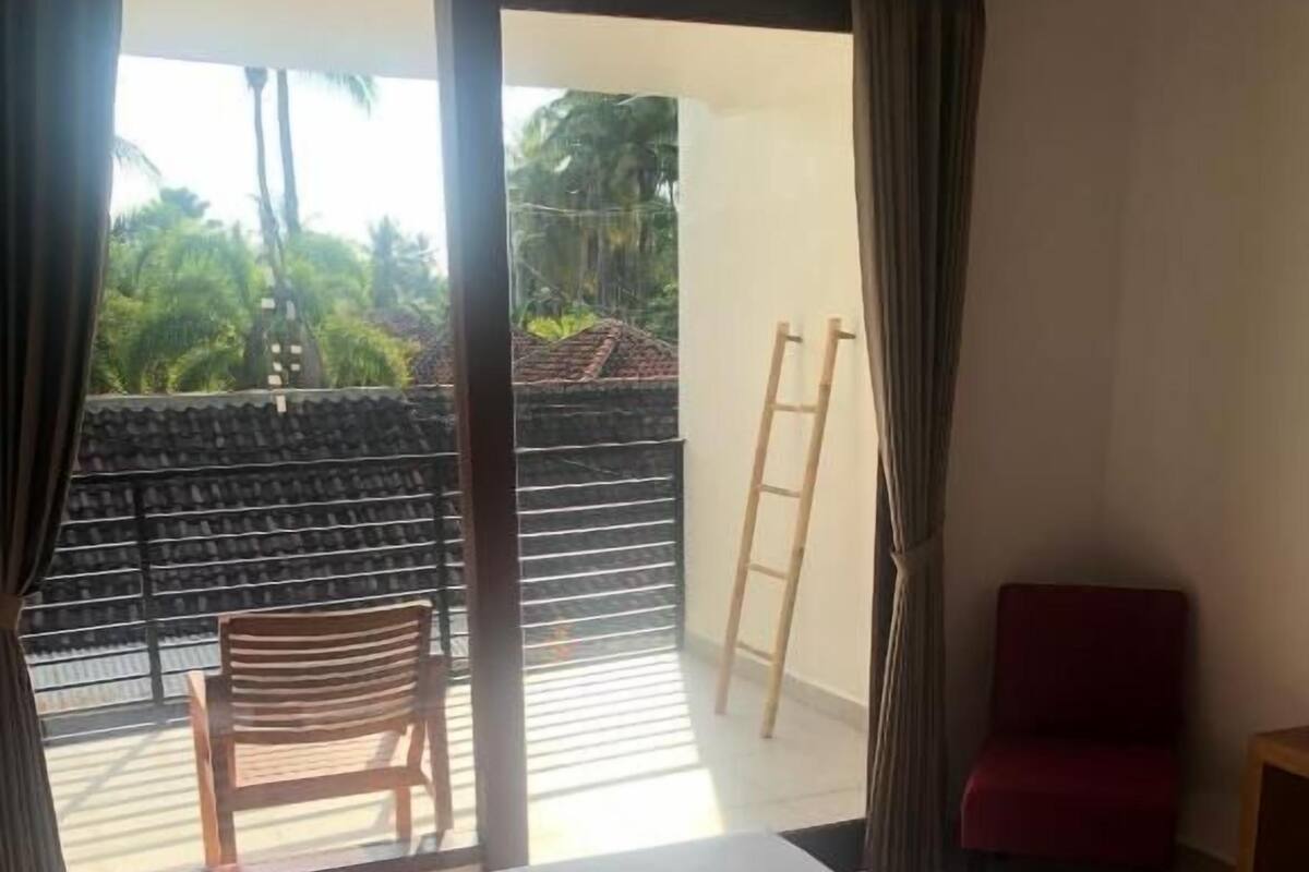Ein Hotelzimmer mit einem Bett, einem roten Sessel, einem Holzstuhl und Blick auf einen Balkon mit Holzgeländer.