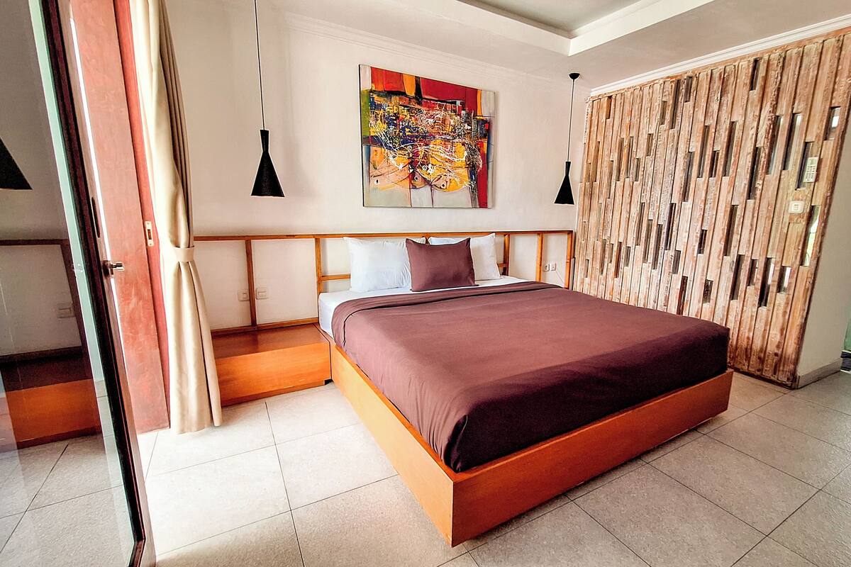 Ein modernes Schlafzimmer mit einem großen Bett, einem hölzernen Kopfteil, einem farbenfrohen abstrakten Gemälde und einer schiebbaren Holztür.