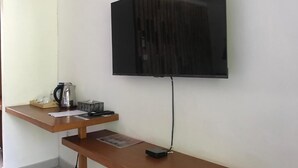 Un televisor de pantalla plana montado en una pared blanca, sobre un escritorio de madera con un hervidor, una caja de pañuelos y un control remoto.