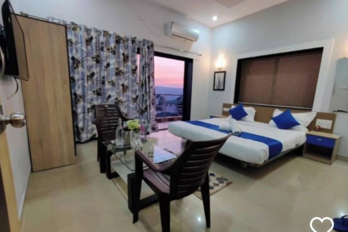 A hotel room with a balcony, a bed with blue bedding, a desk with a chair, and a TV.