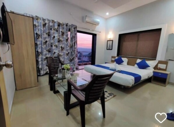 Habitación de hotel con balcón, cama con ropa de cama azul, escritorio con silla y televisor.