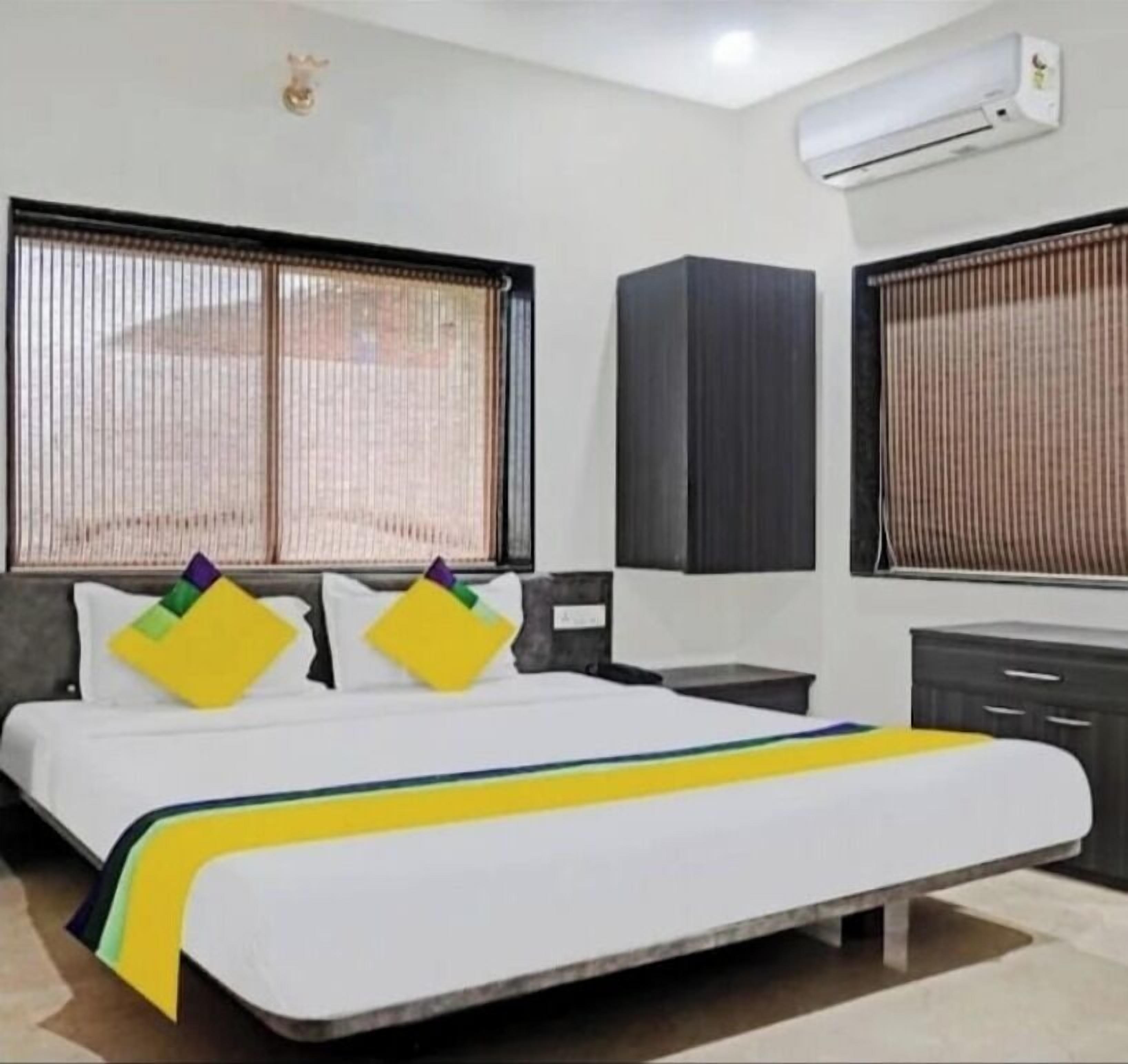 A hotel room with a bed, a desk, a wardrobe, and two windows with blinds.