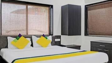 A hotel room with a bed, a desk, a wardrobe, and two windows with blinds.