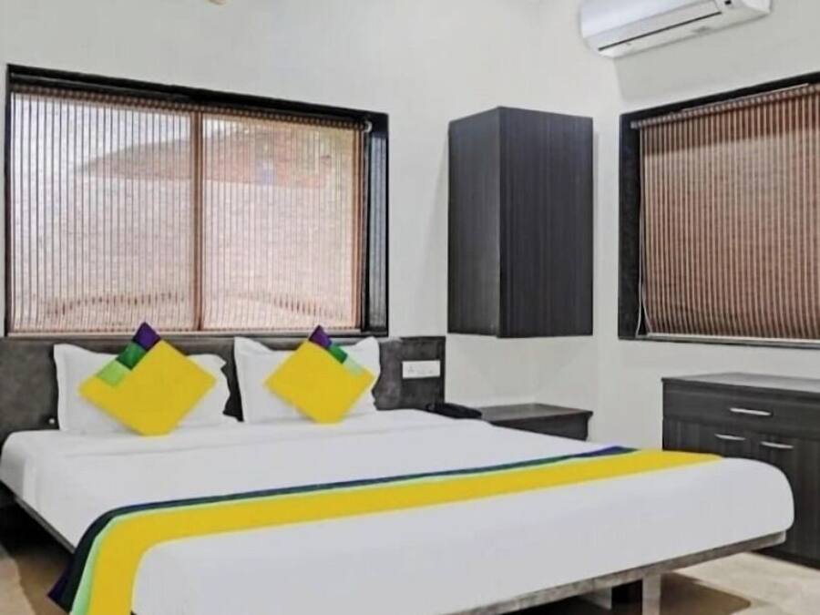 A hotel room with a bed, a desk, a wardrobe, and two windows with blinds.