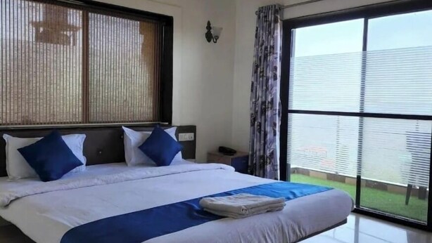 A hotel room with a large bed, a nightstand, an air conditioning unit, and a view of a green area through the window. - The Gren Villa Valley View (Mahabaleshwar)