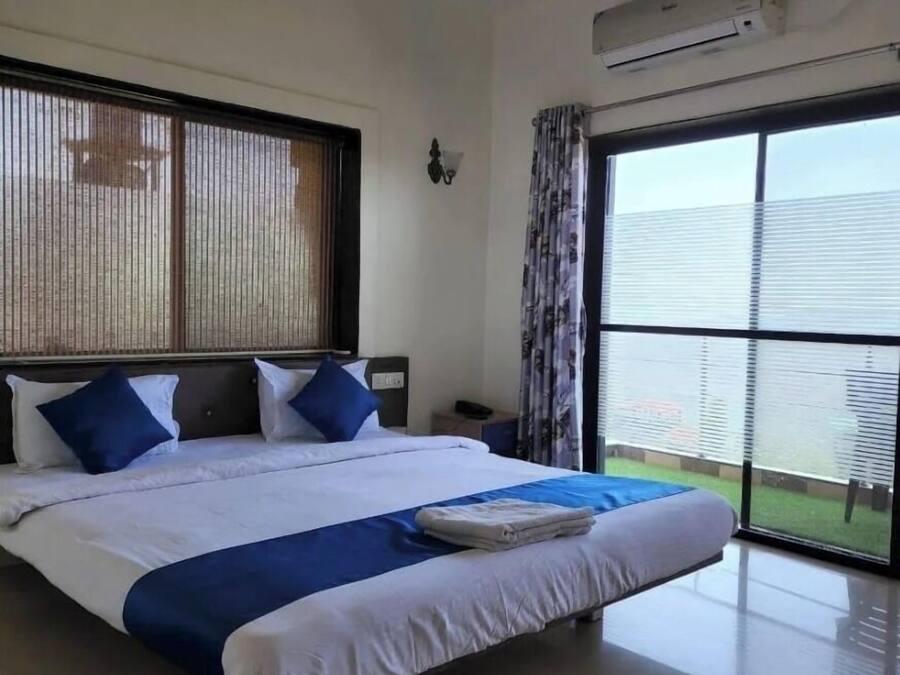 A hotel room with a large bed, a nightstand, an air conditioning unit, and a view of a green area through the window.