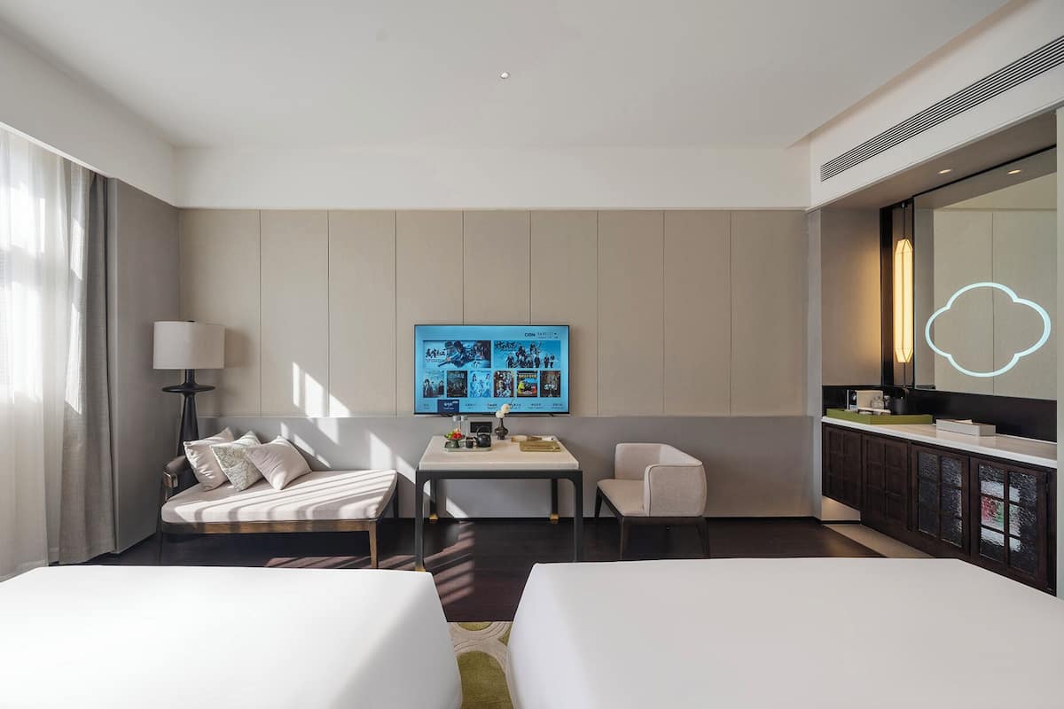 Habitación de hotel con dos camas, un escritorio con lámpara, un televisor mostrando carteles de películas y un espejo grande con luz.