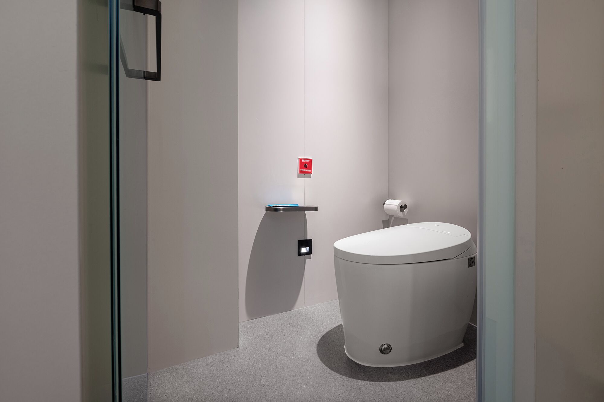 A modern bathroom with a toilet, a small shelf, and a red emergency button on the wall.