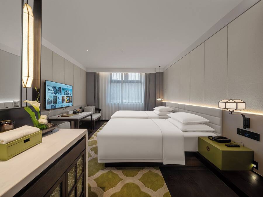 A modern hotel room with a large bed, a desk, a television, and a bedside table with a lamp.