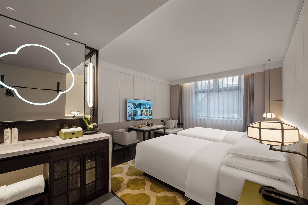 Una habitación de hotel moderna con una cama grande, un escritorio con espejo, un televisor y una mesita de noche con lámpara.