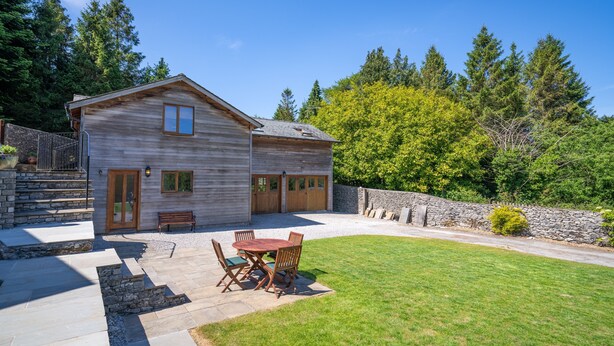 Une maison en bois à deux étages, dotée d’un escalier en pierre, d’une terrasse avec une table et des chaises, et d’une pelouse bien entretenue.