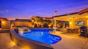 A swimming pool with a curved edge, surrounded by a patio area with outdoor seating and a TV screen mounted on the wall. - Vibrant Home & Sparkling Pool. Entertain in Luxury at this Desert Gem! #75-354 (Bermuda Dunes)