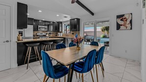 A modern kitchen with dark cabinets, a central island, and a dining area with blue chairs. - Vibrant Home & Sparkling Pool. Entertain in Luxury at this Desert Gem! #75-354 (Bermuda Dunes)