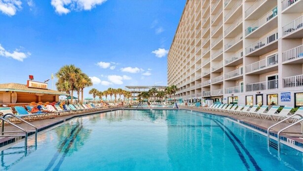 Une piscine d’hôtel avec des chaises longues, un bar et un immeuble de grande hauteur doté de balcons.