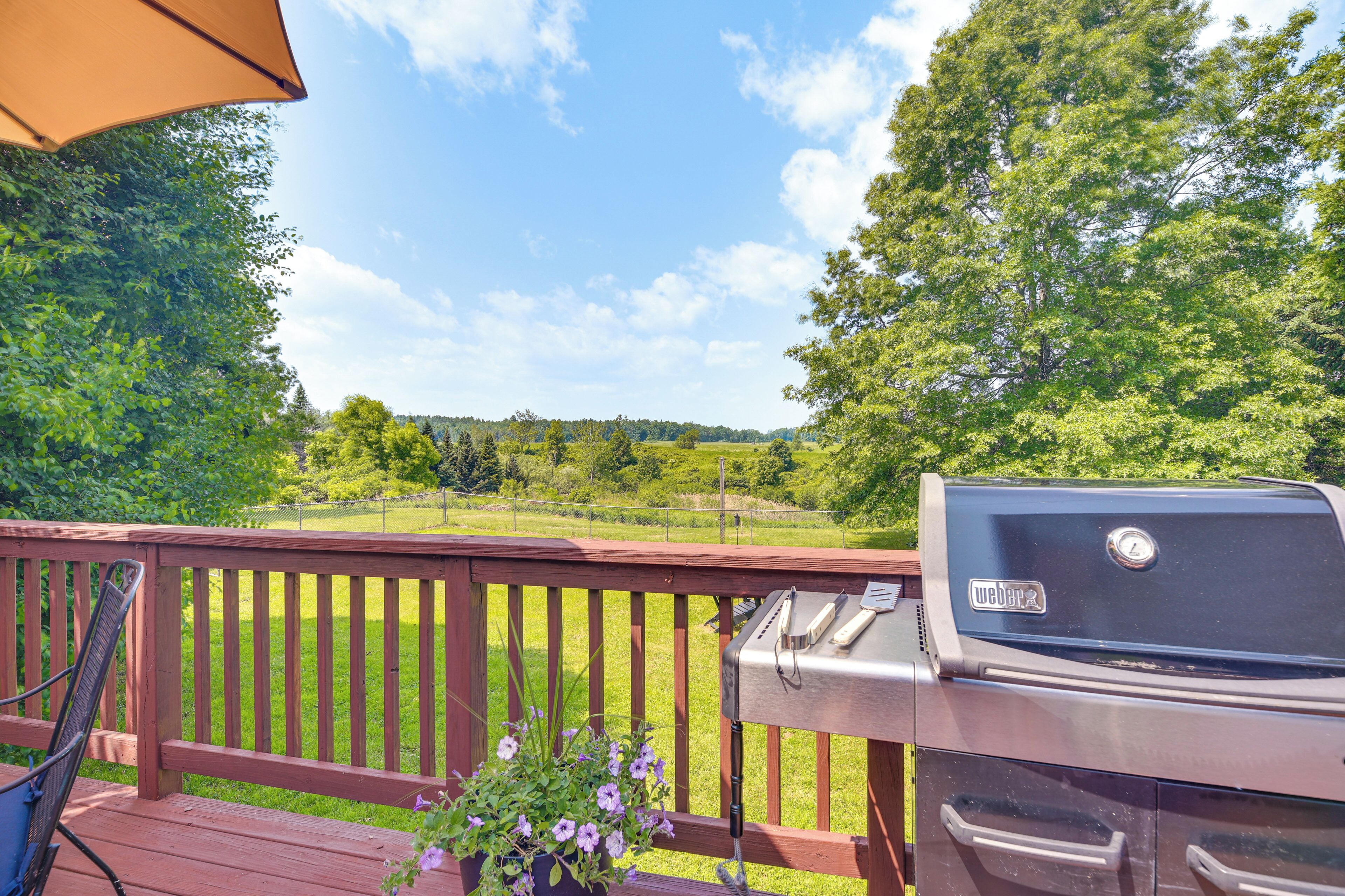 Une terrasse en bois avec un barbecue et une chaise, donnant sur une zone herbeuse bordée d’arbres.