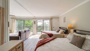 Quarto de hotel com duas camas, um sofá, cômoda de madeira e varanda com vista para árvores.