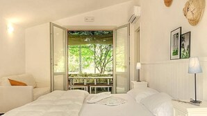 Un dormitorio con dos camas, una ventana con vistas a la vegetación, un sofá blanco y una mesita de noche con una lámpara.