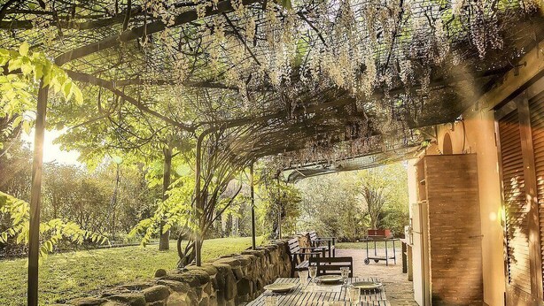 Un área de comedor al aire libre con mesas y sillas, rodeada de muros de piedra y follaje.