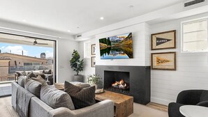 A modern living room with a fireplace, a large TV, and a view of the city. - 3BR + Loft Ocean View | Rooftop, Hot Tub, Parking (Newport Beach)