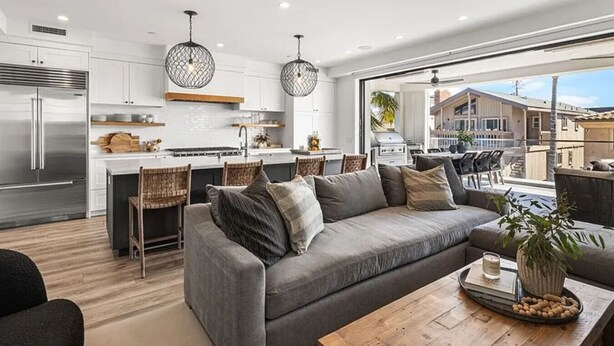 A modern kitchen with a central island, a dining area with chairs, a grey sofa, and a large window overlooking a residential area. - 3BR + Loft Ocean View | Rooftop, Hot Tub, Parking (Newport Beach)