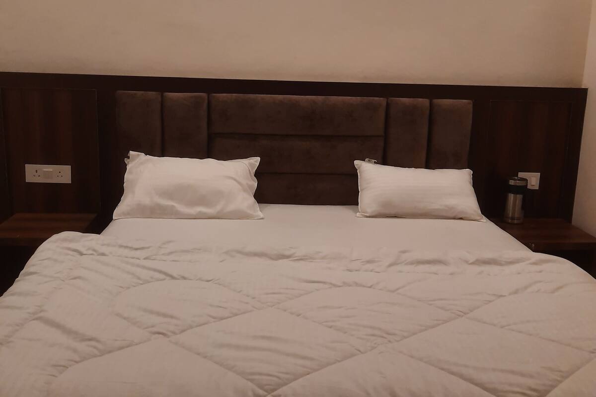 A hotel room with a large bed, two white pillows, a wooden headboard, and a bedside table with a lamp.