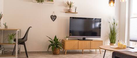 Een woonkamer met een flatscreen televisie op een houten kast, een eettafel met stoelen en verschillende potplanten.