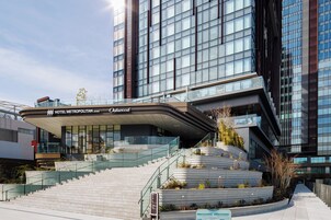A modern hotel entrance with a curved glass facade, a staircase leading to the entrance, and a landscaped area with plants.