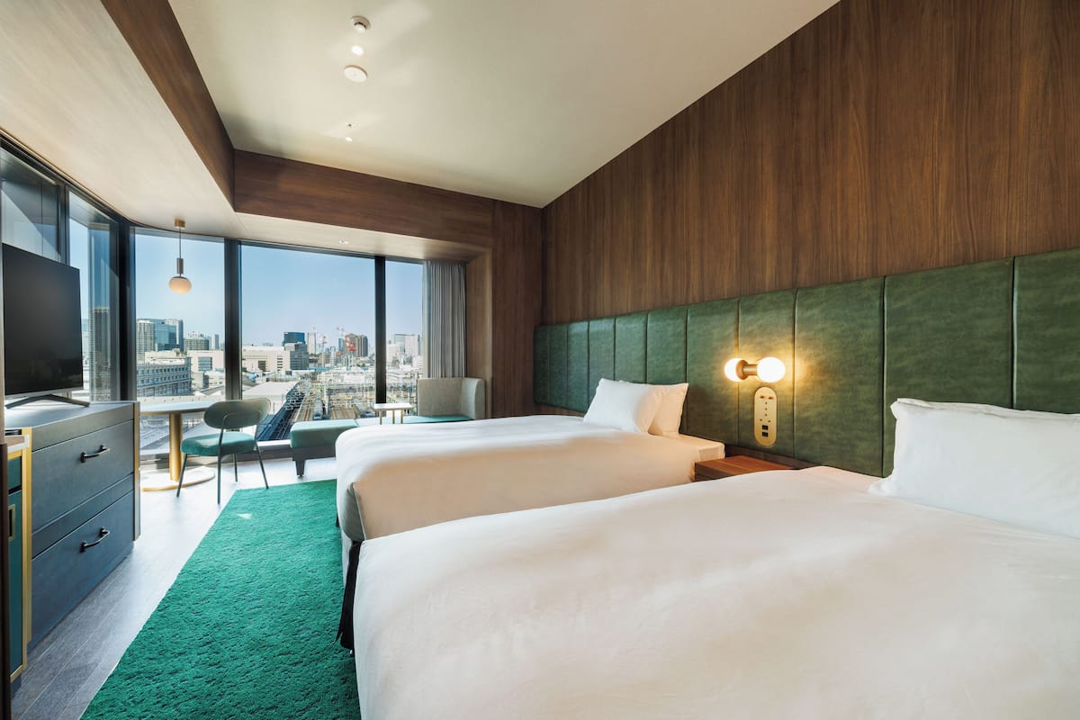 A hotel room with two beds, a desk, a chair, and a large window offering a city view.