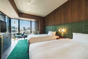A hotel room with two beds, a desk, a chair, and a large window offering a city view.