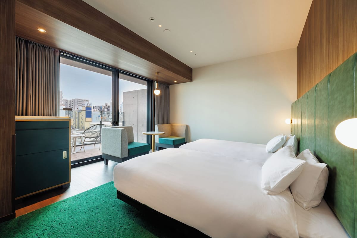 A modern hotel room with a large bed, a green carpet, a desk, and a view of the city through large windows.