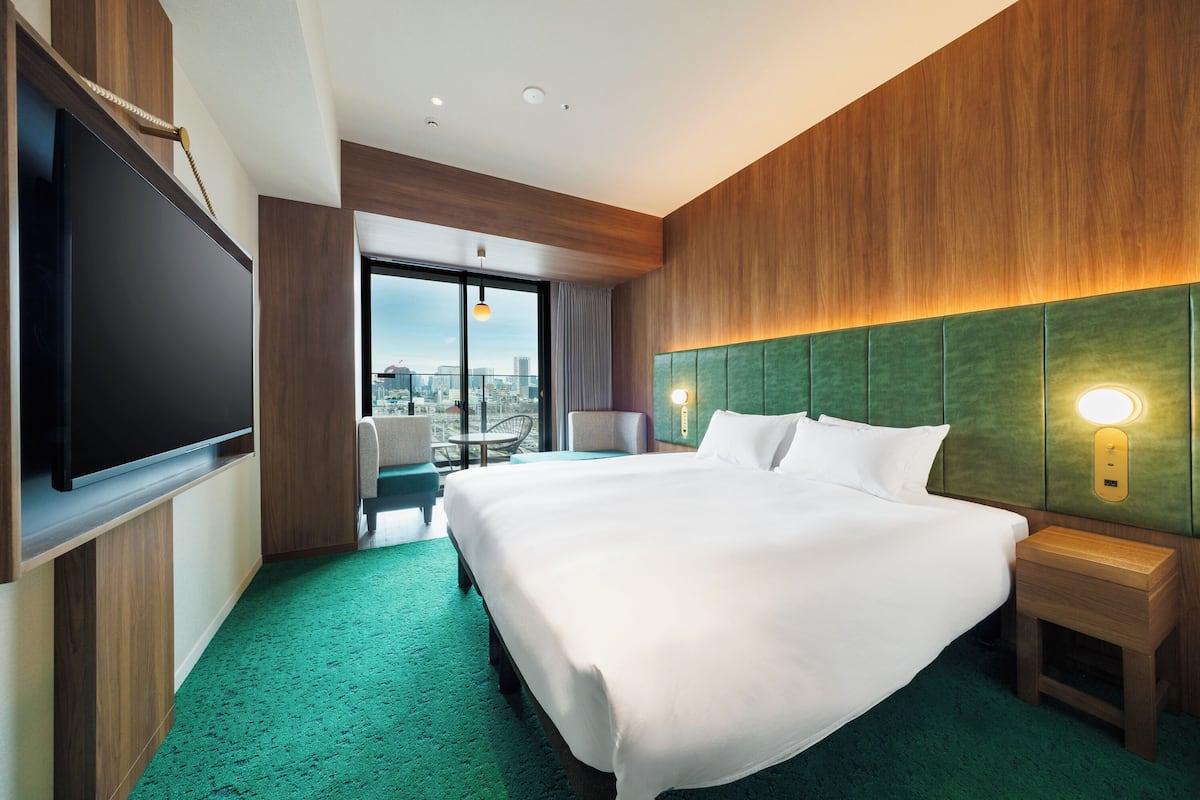 A hotel room with a large bed, a flat-screen TV, a green upholstered headboard, and a city view.