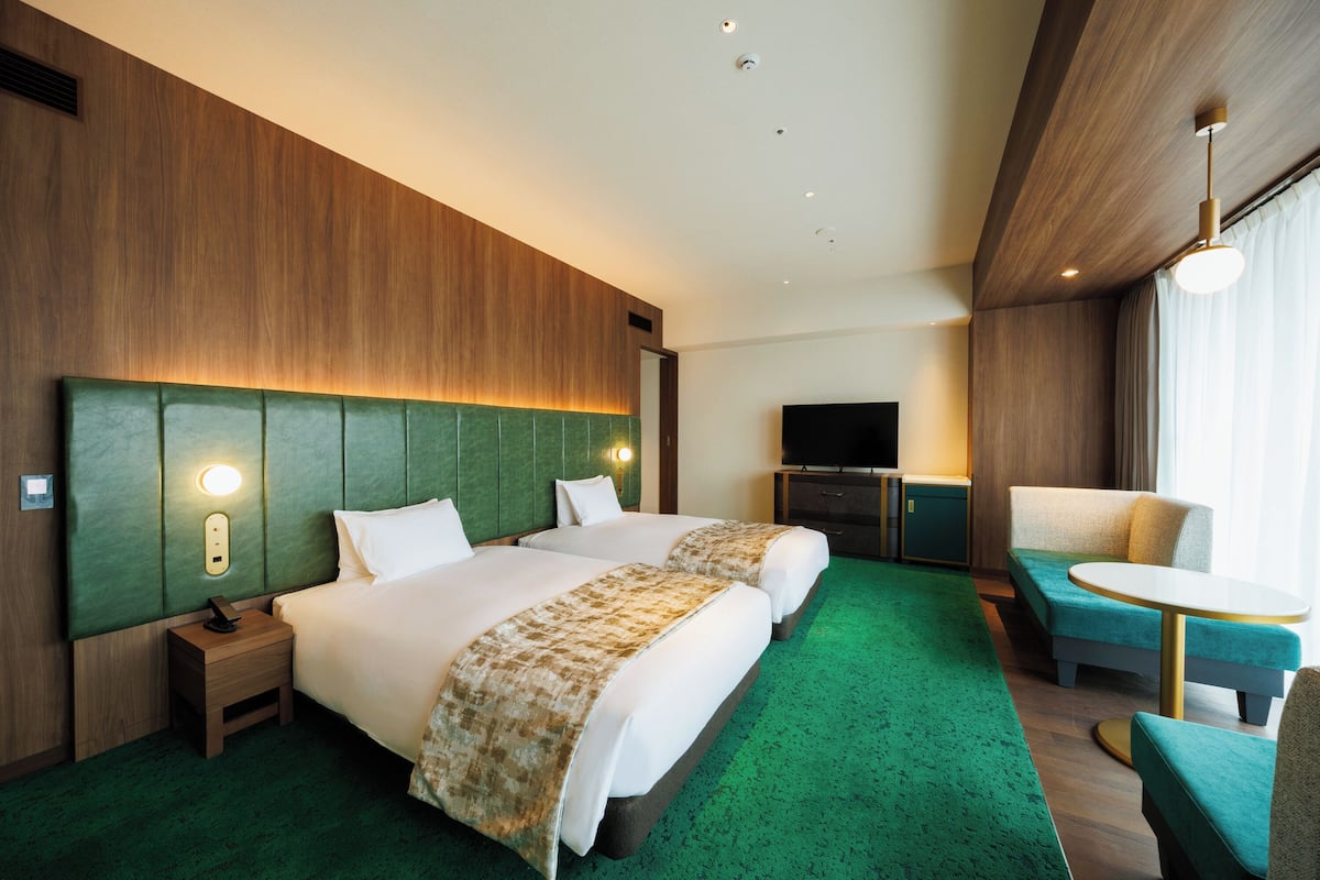 A hotel room with two beds, a TV, a small table, and a green carpet.