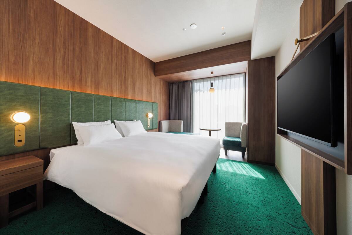 A hotel room with a large bed, a wooden headboard, a flat-screen TV, a desk with a lamp, and a view of the outdoors through a window.