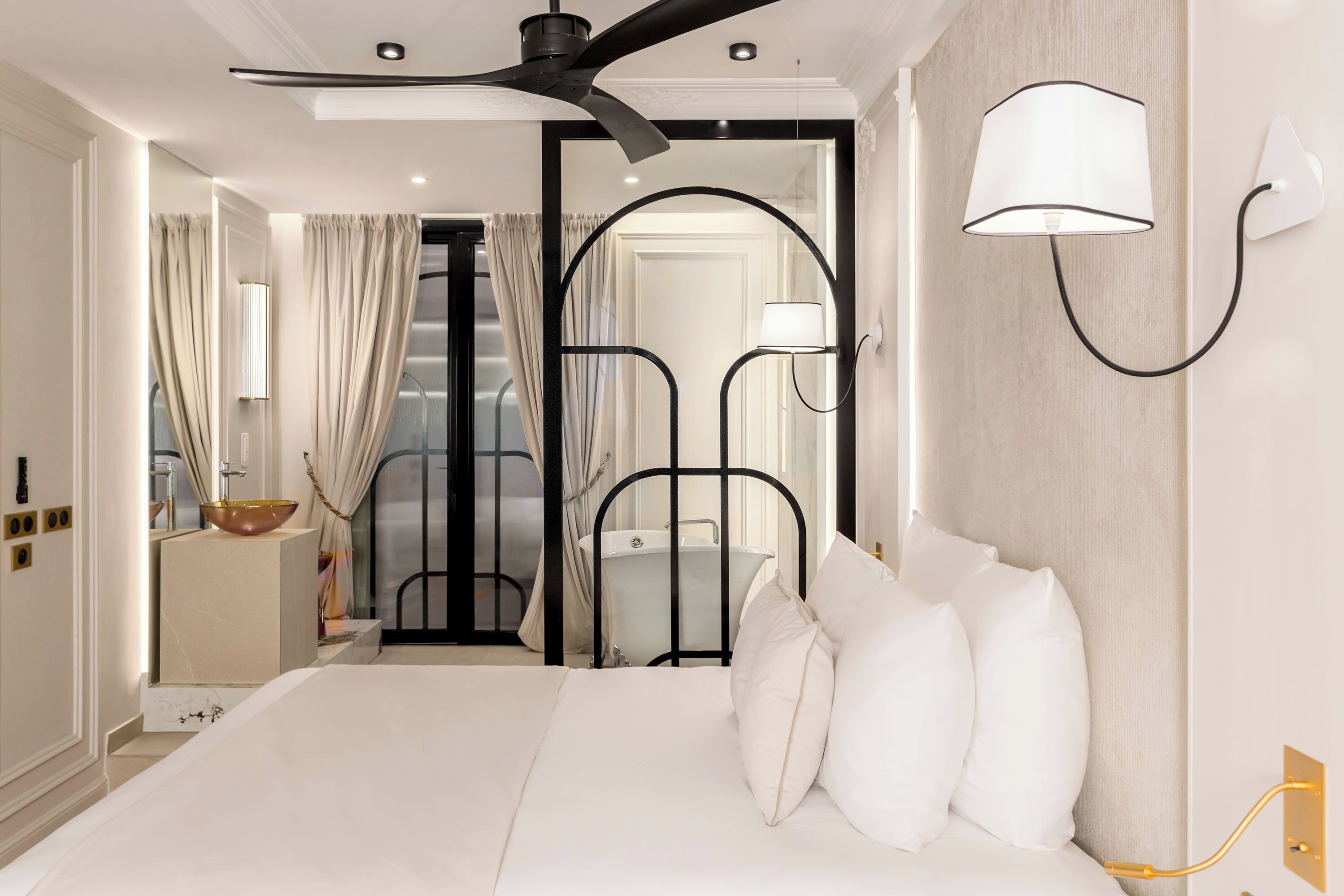 A modern hotel room with a large bed, a ceiling fan, a wall-mounted lamp, and a view of the bathroom through an open door.