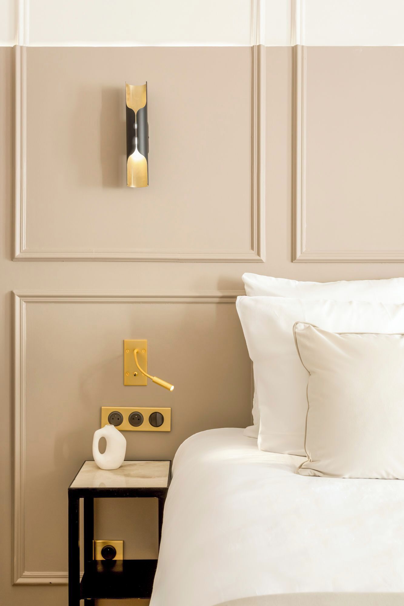 A hotel room with a bed, a nightstand with a lamp, and a wall-mounted power outlet.