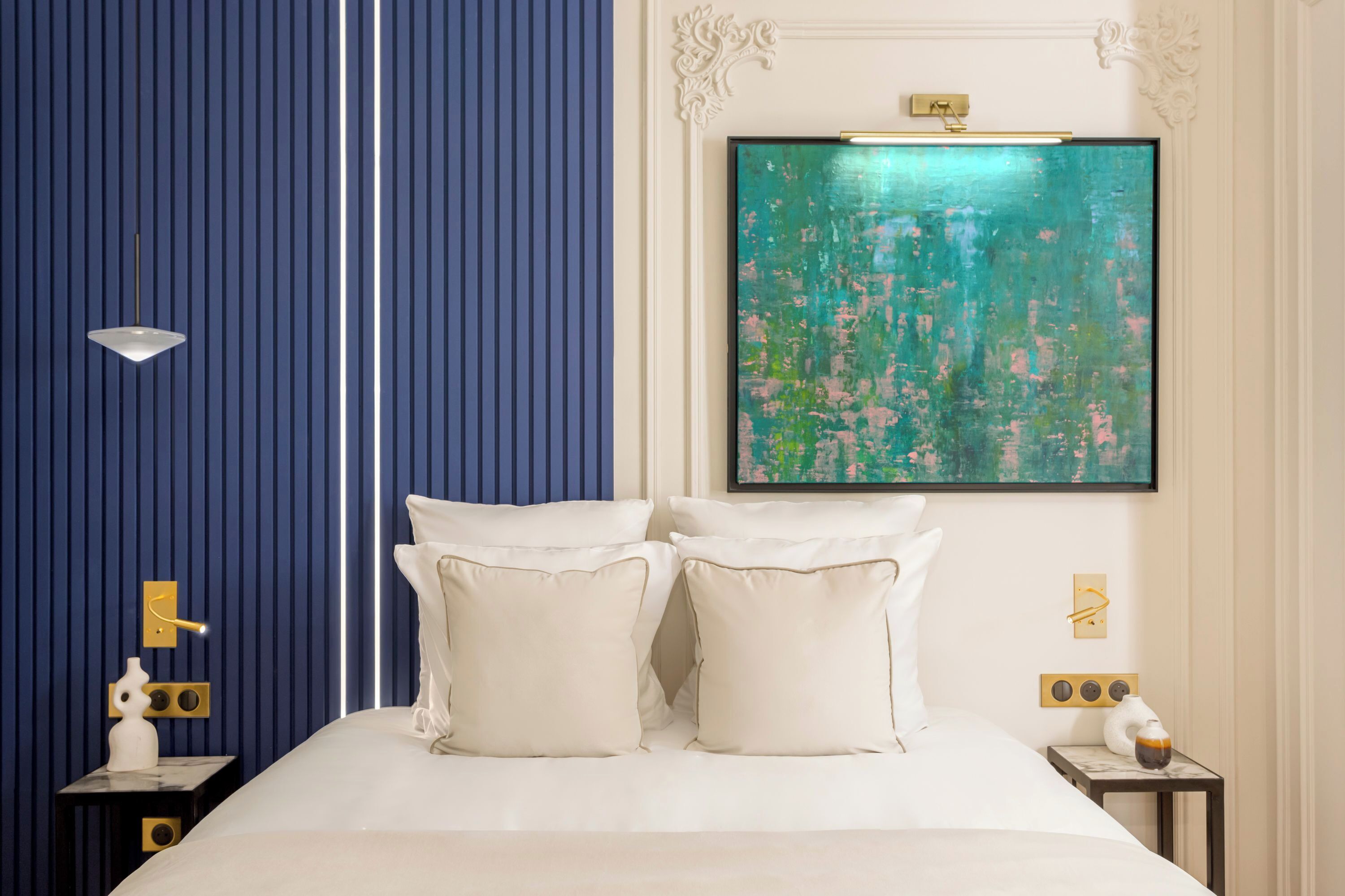 Camera d'albergo con un letto, due cuscini bianchi, un comodino con una lampada, un tavolino con una teiera e un grande quadro astratto appeso alla parete.
