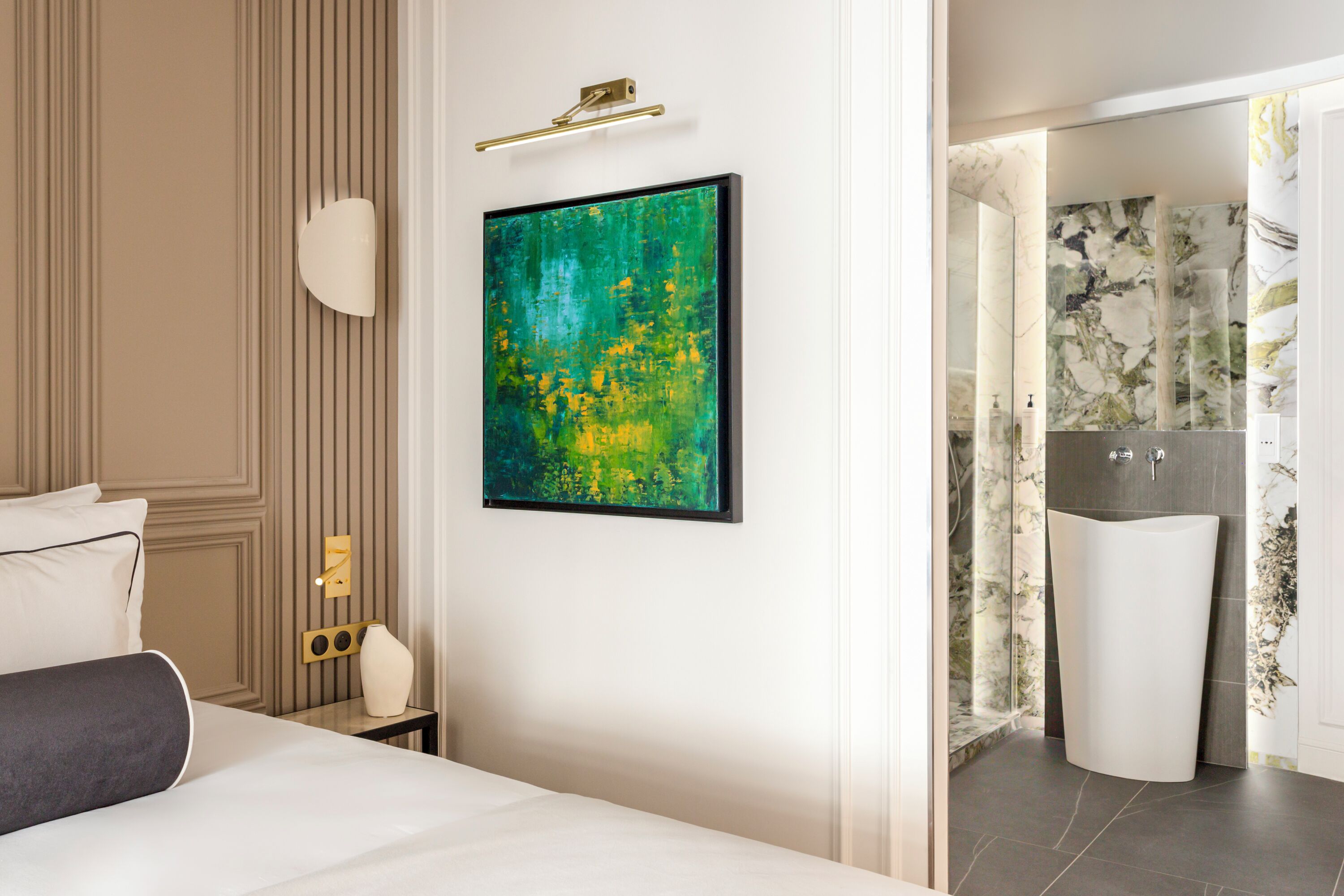 A modern hotel room with a bed, a nightstand, a wall-mounted light, and a bathroom with a sink and mirror.