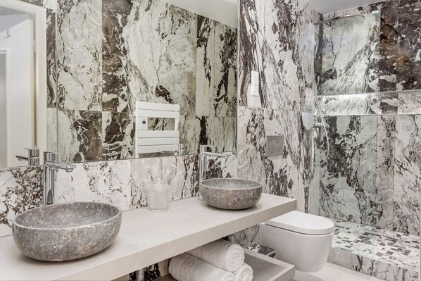 Un baño con paredes de mármol, dos lavamanos, un inodoro y una ducha.