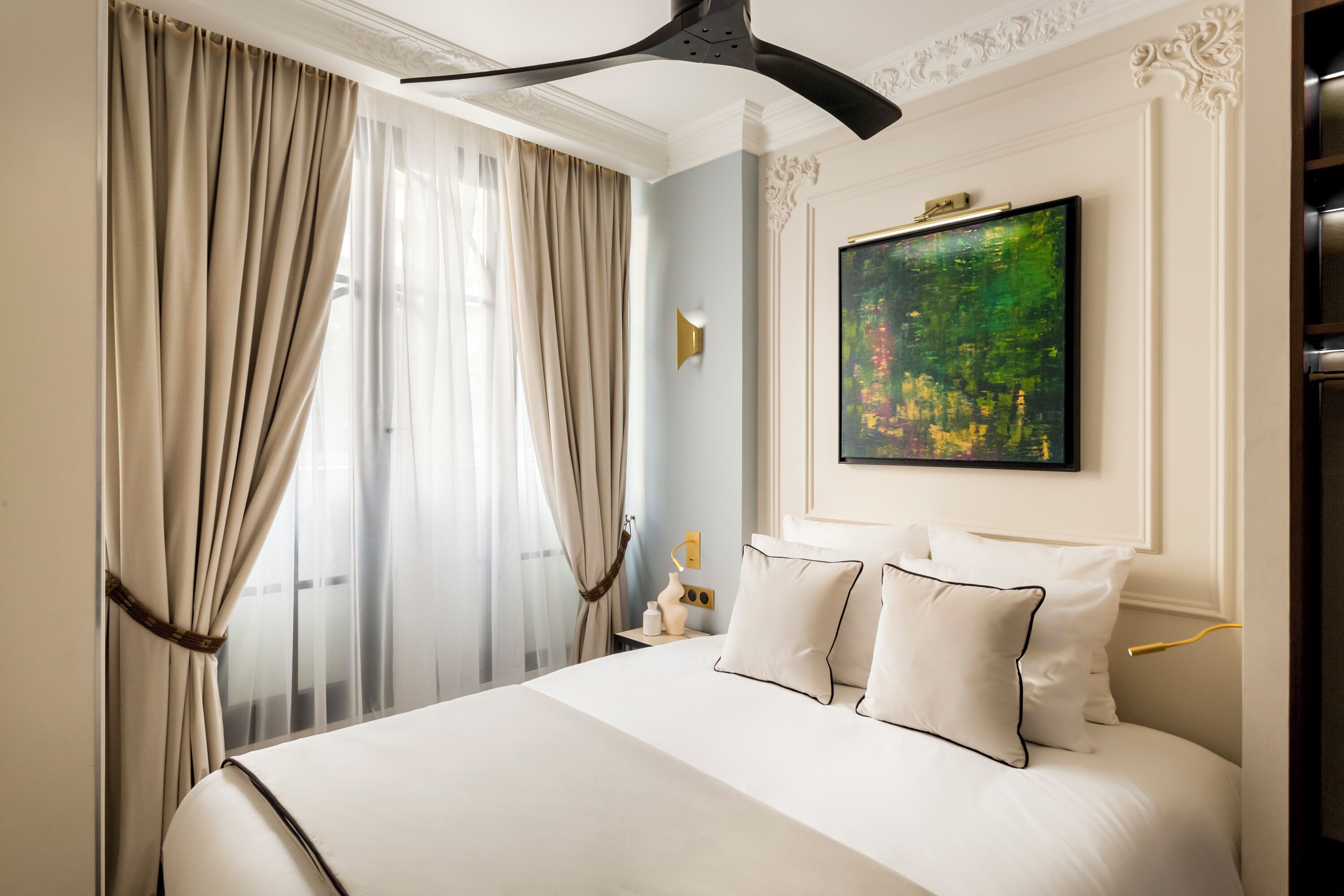 Ein Hotelzimmer mit einem großen Bett, beigen Vorhängen, einem Bild an der Wand und einem Deckenventilator.