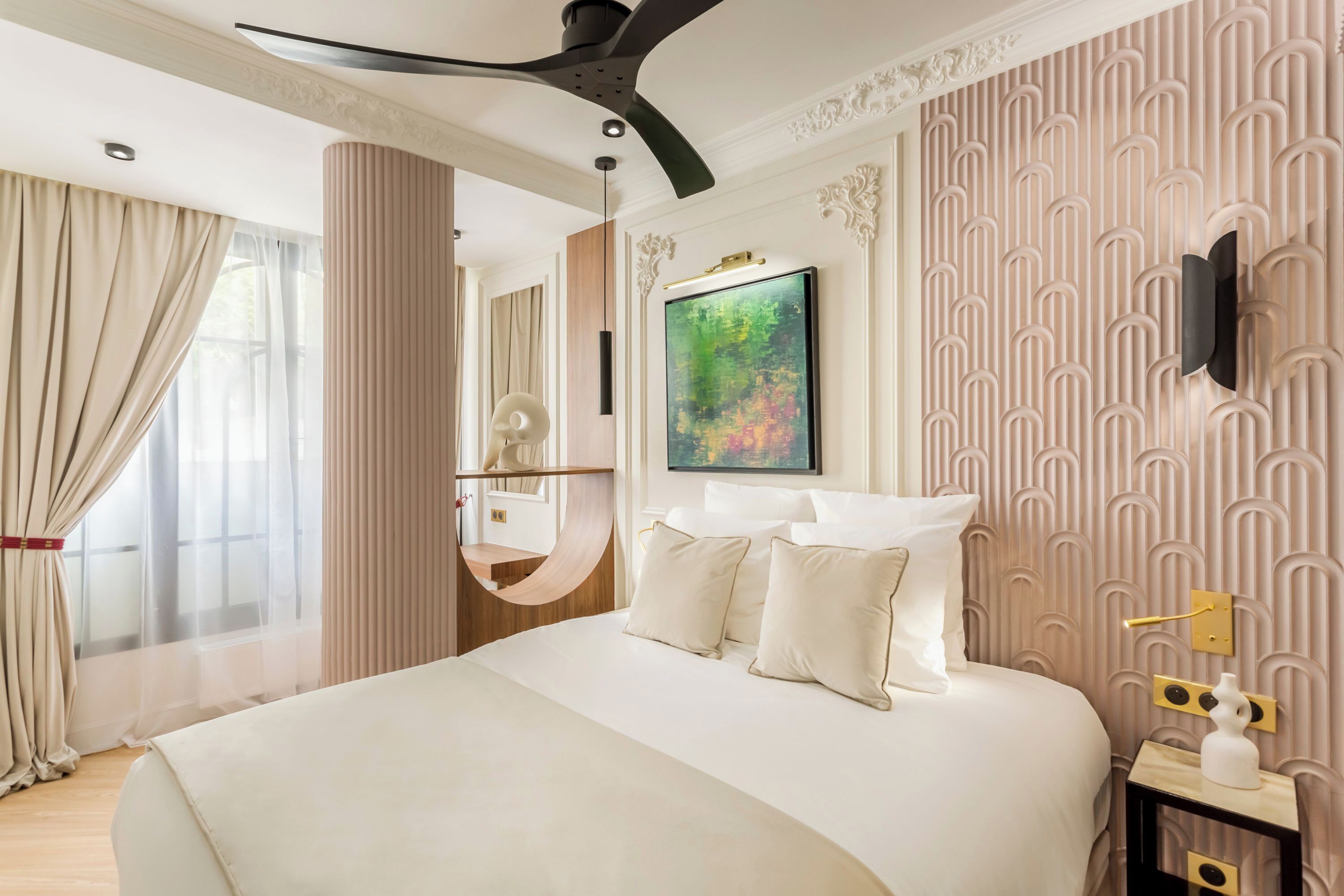 A modern hotel room with a large bed, a ceiling fan, a painting on the wall, and a decorative wall panel.