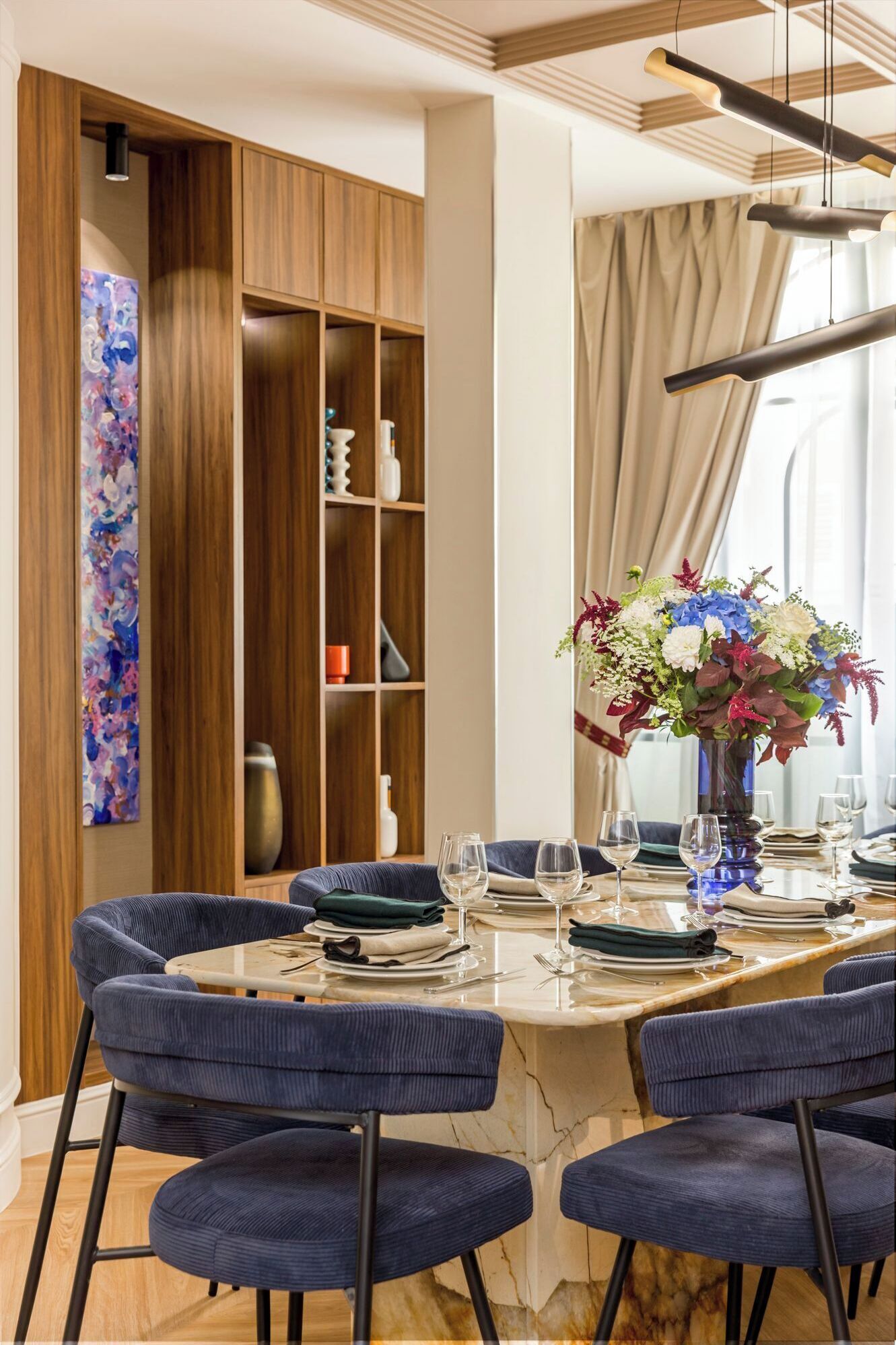 Ein Essbereich mit einem Marmortisch, blau gepolsterten Stühlen, einer Blumenvase und Holzregalen mit Dekoration.