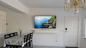 En matplats med ett bord dukat för fyra personer, en väggmonterad tv som visar ett vackert landskap och en kristallkrona ovanför.