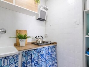 A kitchen with white tiled walls, a wooden countertop, a kettle, a cutting board, and a potted plant.