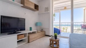 Ein modernes Hotelzimmer mit einem Flachbildfernseher, Holzböden, einem kleinen Beistelltisch mit Pflanze und einem Balkon mit Blick auf den Strand.